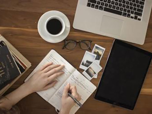 Overhead view of a desk with a laptop, coffee, and hands writing in a book icon