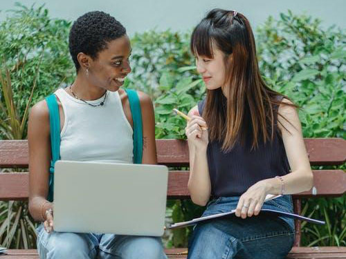 Two women sitting together on a bench outdoor with a laptop and a book and having a conversation icon