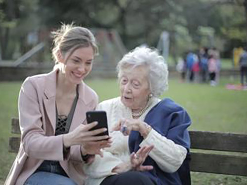 An old and young woman sitting on a bench and looking at a phone together icon